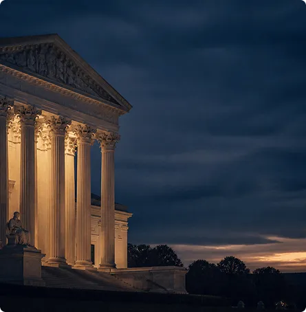 Illuminated neoclassical building facade with Corinthian columns at dusk, dark cloudy sky.