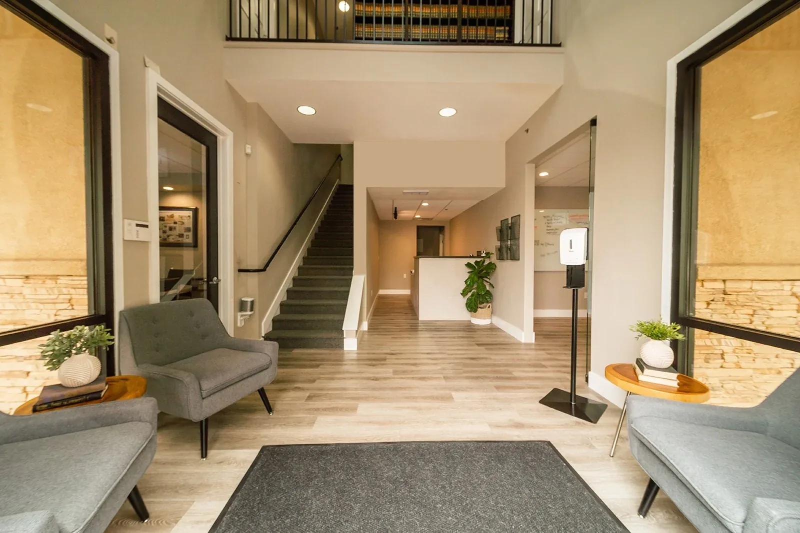Modern office lobby with gray chairs, wooden side tables, indoor plants, and a reception desk.