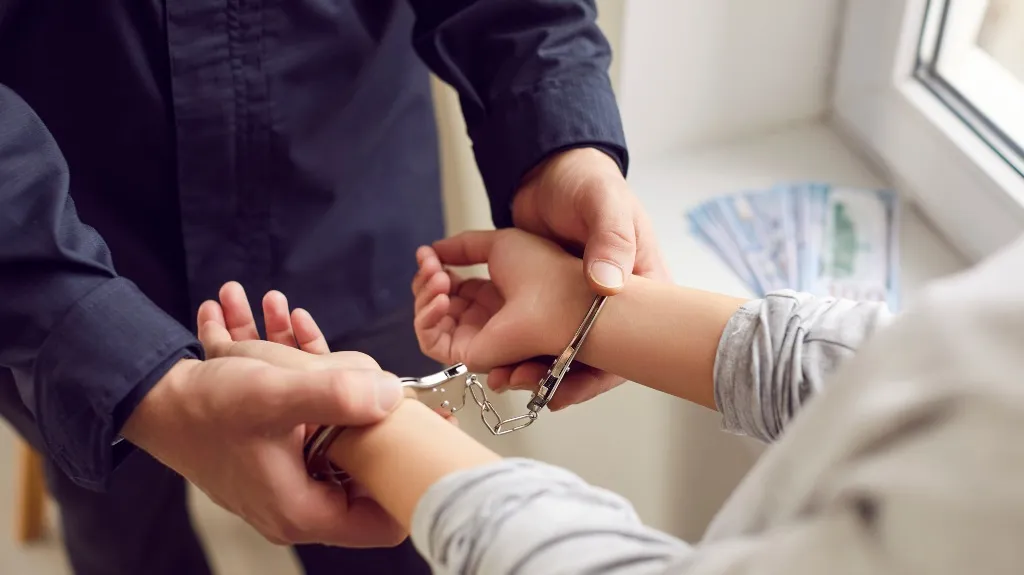 A person in a dark shirt handcuffing another individual near a window with money.