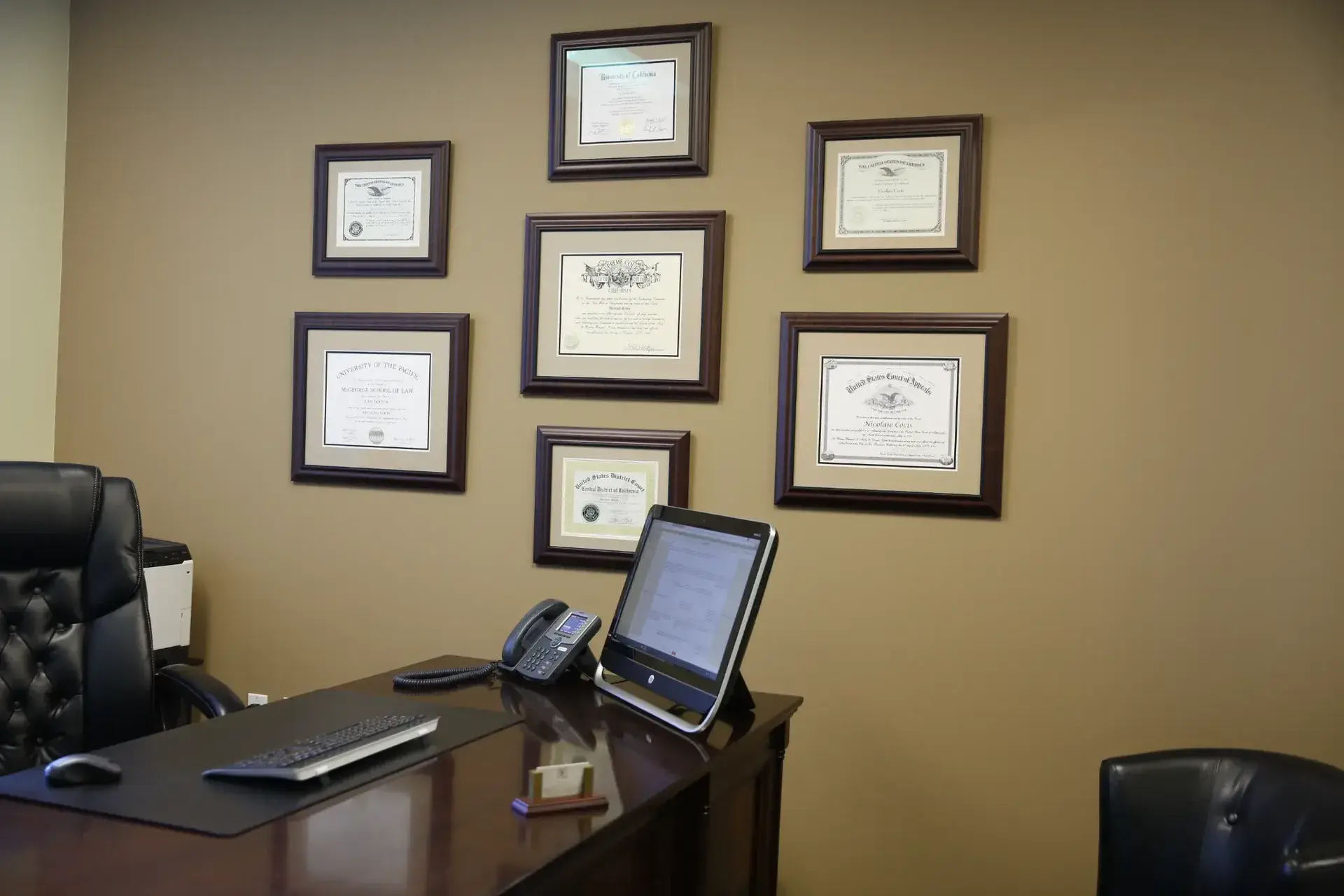 Professional office interior with dark wooden desk, black leather chair, computer, phone, and framed certificates on beige wall.