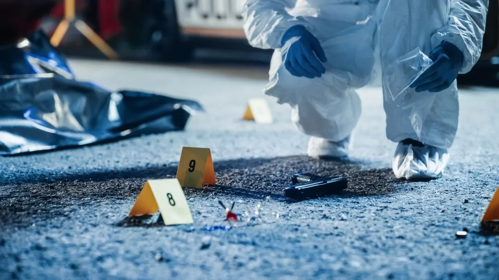 Forensic investigator in white protective suit examining a handgun at a crime scene with evidence markers.