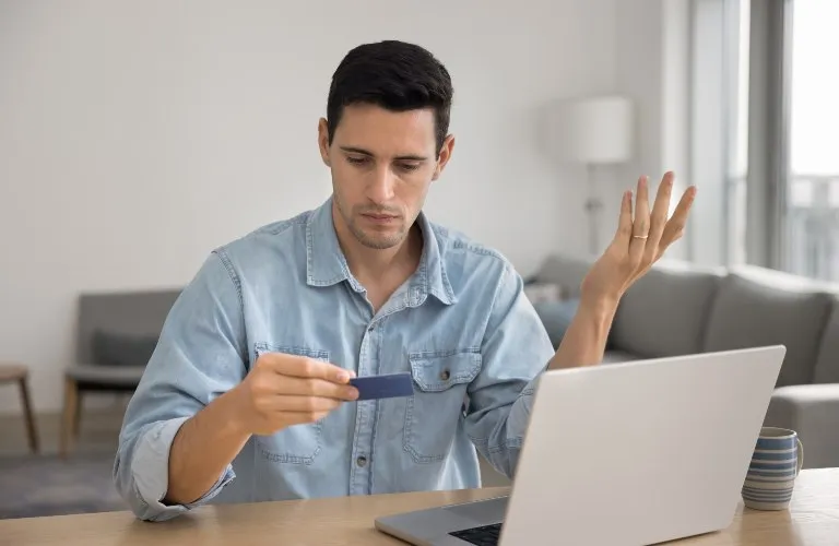 Man in a light blue shirt holding a credit card and looking confused at a laptop.