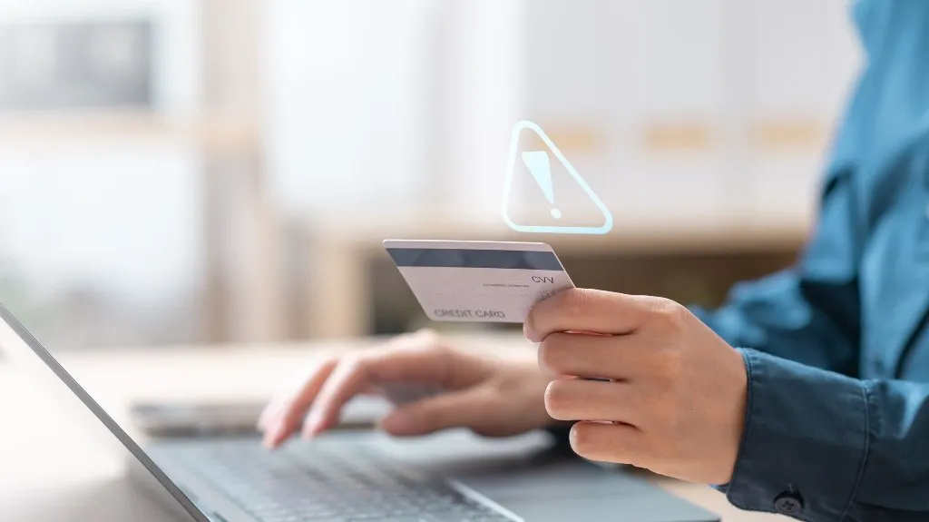 Person holding a credit card near a laptop keyboard with a digital warning icon above.