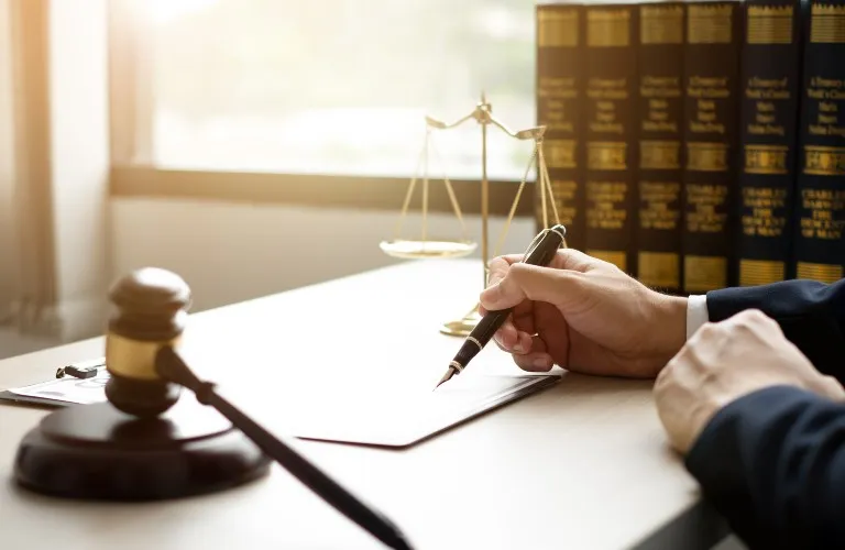 Hand holding pen poised over document on desk with gavel, scales of justice, and legal books.