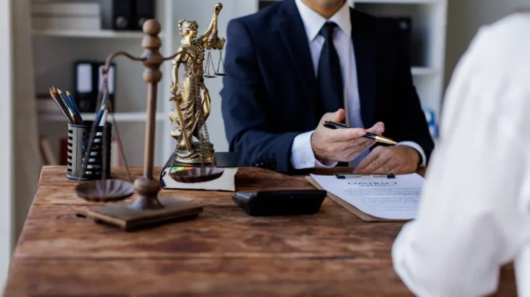 Man in suit holding pen over contract document at wooden desk with legal scales and Lady Justice statue.