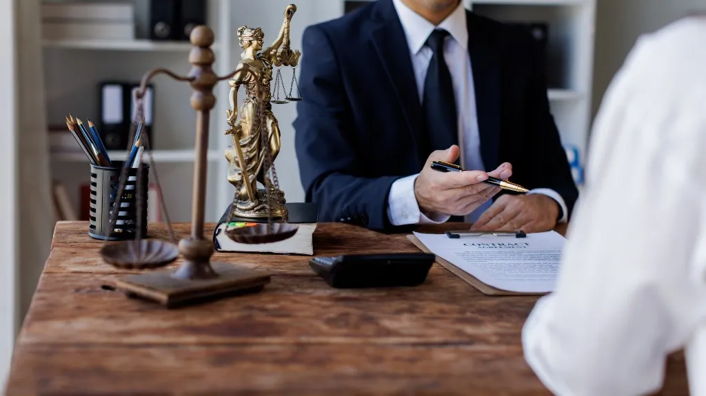 Man in suit holding pen over contract document at wooden desk with legal scales and Lady Justice statue.