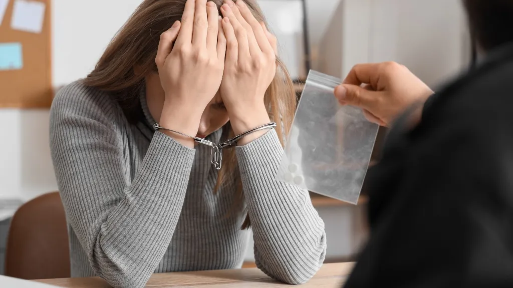 Handcuffed woman in gray sweater covering face, person holding small plastic bag with pills.