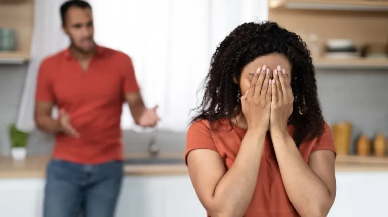A woman covering her face with hands in distress, a man in red shirt gesturing behind her.