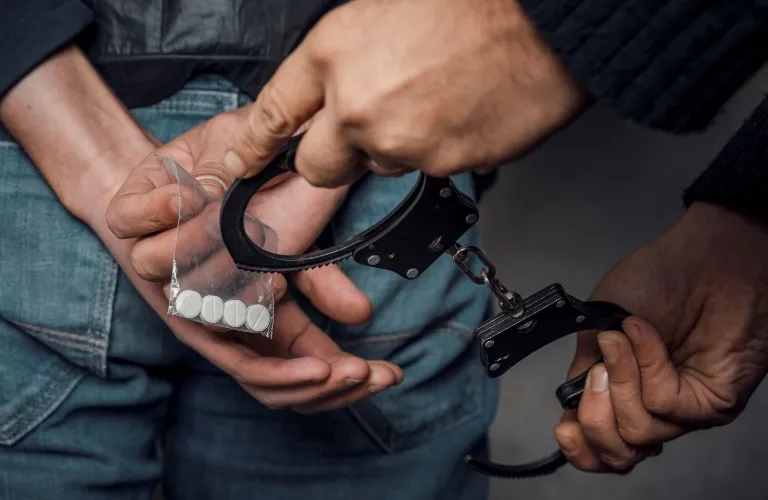 Close-up of a person in blue jeans holding a small bag of white pills, with another person placing black handcuffs.