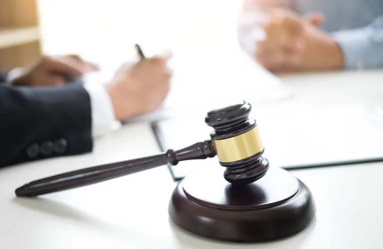 Dark wooden judge's gavel with gold band on white table, blurred people in background.