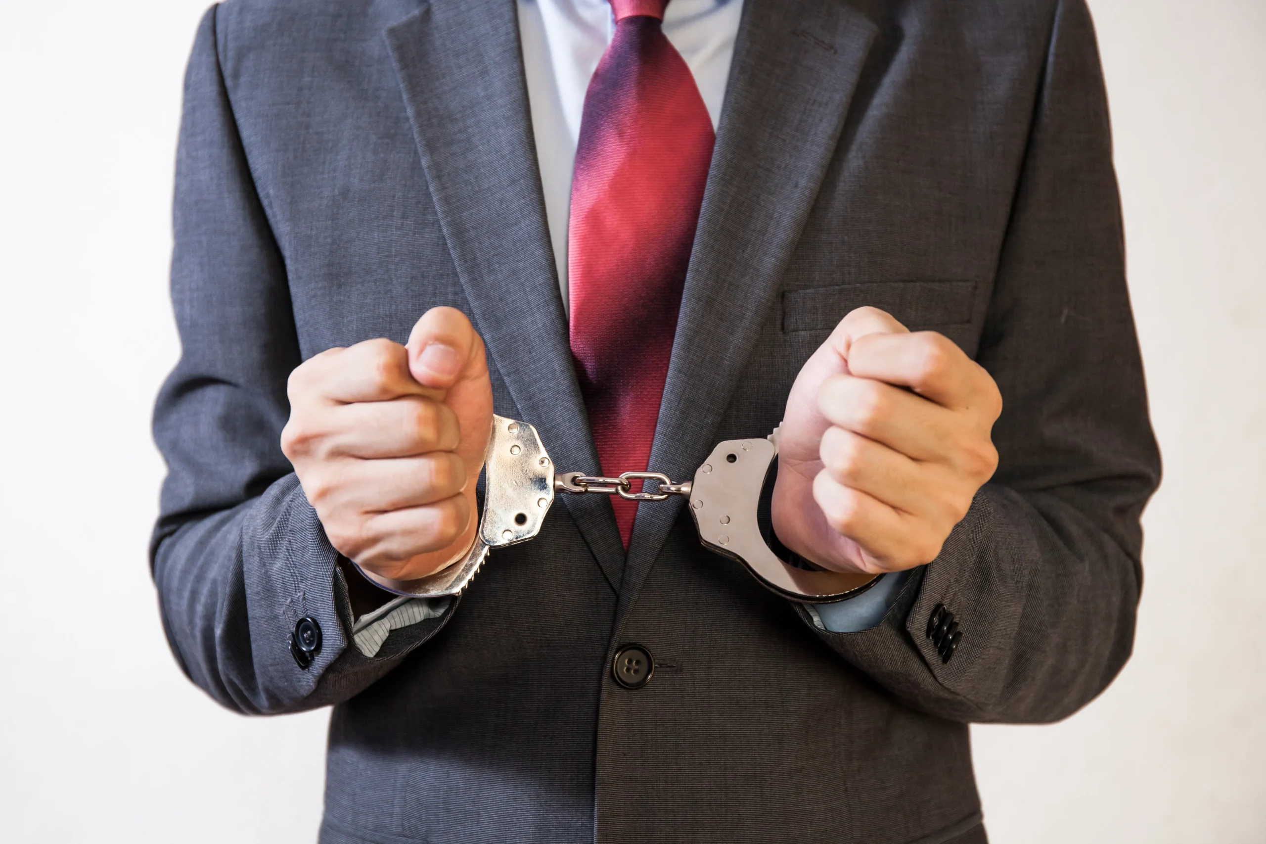 Man in gray suit and red tie wearing metal handcuffs on wrists.