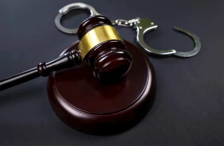 Wooden judge's gavel with brass band resting on sound block next to metal handcuffs.