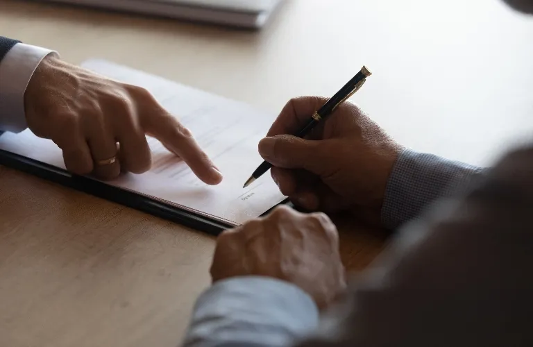 Two individuals reviewing and signing a document on a wooden table, one pointing while the other holds a pen.