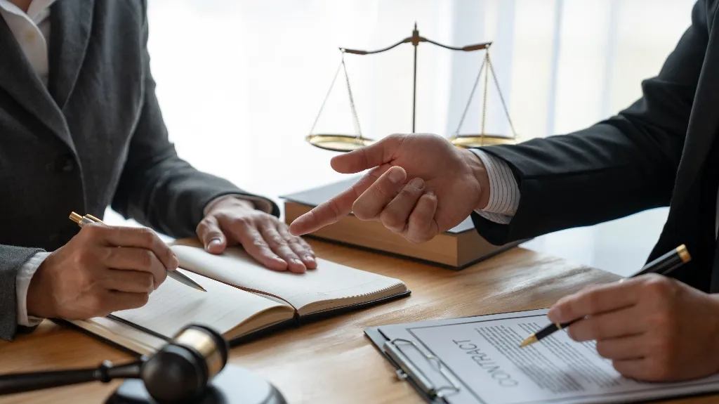 Two professionals in suits discussing and signing a contract with legal scales and gavel on the desk.